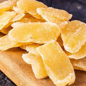 May include: A close-up shot of candied ginger slices, showcasing their golden-yellow hue and crystallized sugar coating. The ginger pieces are arranged on a wooden cutting board, creating a visually appealing display of the sweet treat.