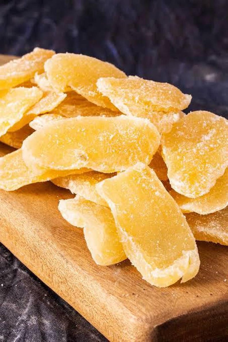 May include: A close-up shot of candied ginger slices, showcasing their golden-yellow hue and crystallized sugar coating. The ginger pieces are arranged on a wooden cutting board, creating a visually appealing display of the sweet treat.