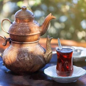 May include: A copper tea set, including a teapot and a glass of tea on a white saucer. The teapot features detailed patterns and a curved spout. The tea is a deep red hue, with a spoon in the glass. The set is presented on a dark wooden tray.