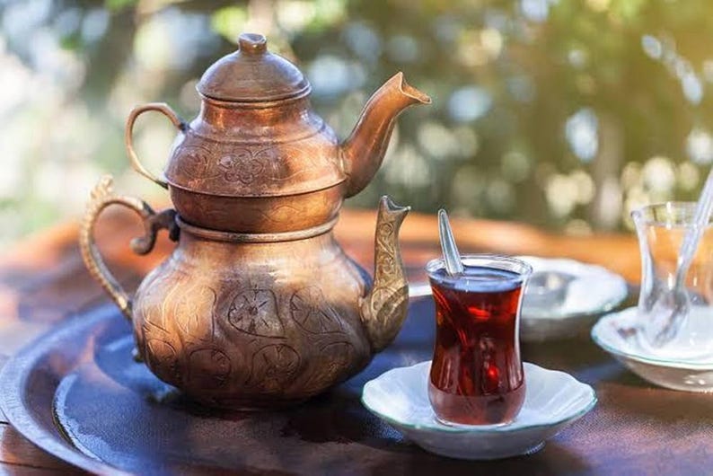 May include: A copper tea set, including a teapot and a glass of tea on a white saucer. The teapot features detailed patterns and a curved spout. The tea is a deep red hue, with a spoon in the glass. The set is presented on a dark wooden tray.