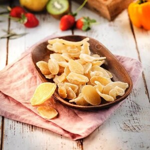 May include: A wooden bowl filled with candied ginger slices. The ginger is a light amber color and has a crystallized sugar coating. The bowl sits on a pink cloth, with strawberries and kiwi in the background.