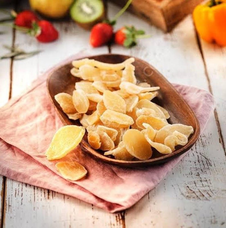May include: A wooden bowl filled with candied ginger slices. The ginger is a light amber color and has a crystallized sugar coating. The bowl sits on a pink cloth, with strawberries and kiwi in the background.