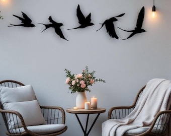 Puede incluir: Decoración de pared con cinco siluetas de pájaros negros en vuelo. Los pájaros están montados en una pared blanca sobre dos sillas de mimbre, una mesa pequeña y un jarrón con flores. Luces de cadena cuelgan por encima.