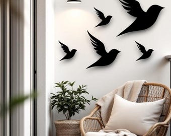 Puede incluir: Decoración mural de pájaros negros. Seis siluetas de pájaros negros de varios tamaños están montadas en una pared blanca. Debajo de la decoración de la pared hay una silla de mimbre con un cojín blanco y una manta. A la izquierda hay una planta en maceta.