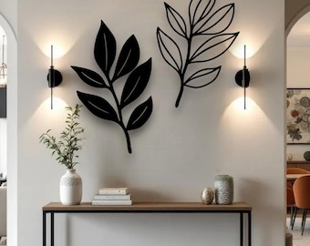Puede incluir: Dos piezas de arte mural de hojas de metal negro. Un diseño de hoja es sólido, el otro es un contorno. Las hojas están montadas en una pared blanca, con apliques a ambos lados. Debajo hay una consola de madera con marco de metal negro.