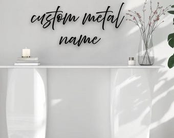 May include: Black metal sign with the words "custom metal name" in a cursive font. The sign is mounted on a white wall above a white console table. A vase with flowers and a candle are on the table.