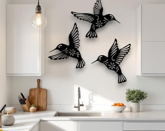 Puede incluir: Tres piezas de arte mural de metal negro con forma de colibríes. Cada ave se representa en vuelo con alas y plumas de la cola detalladas. Los colibríes tienen detalles florales y están montados en una pared blanca.