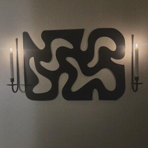 K&ouml;nnte beinhalten: Eine schwarze Wandskulptur mit einem durchgehenden, flie&szlig;enden, abstrakten Ausschnittmuster. Die Skulptur wird von zwei vertikalen Metallkerzenhaltern flankiert, die jeweils eine brennende wei&szlig;e Stabkerze halten und einen warmen Schein erzeugen.