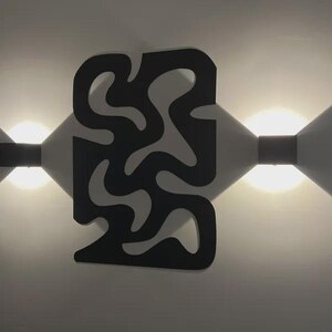 K&ouml;nnte beinhalten: Eine schwarze Wandleuchte mit einer zentralen Platte, die ein ausgeschnittenes, abstraktes Wellenmuster aufweist. Die Leuchte ist zwischen zwei hellen Lichtquellen positioniert, die dreieckige Schatten an die Wand werfen und die dunkle Form betonen.