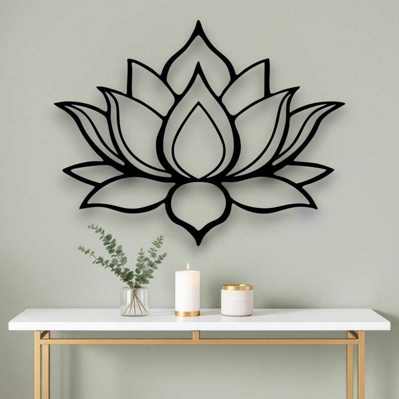 May include: A black lotus flower wall art decor. The metal art piece is mounted on a light green wall. Below the art is a white table with gold legs, a candle, and a small vase with greenery.