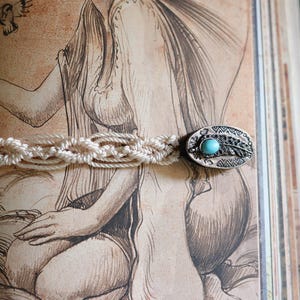 May include: A macrame bracelet with an oval silver-tone clasp featuring a turquoise stone and feather design. The bracelet is set against a vintage illustration background, with a detailed woven pattern.