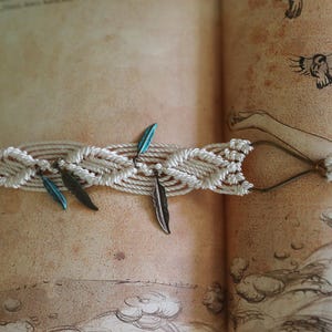 May include: A cream-colored macrame headband with three decorative feathers. Two feathers are teal, and one is silver. The headband is displayed on an open book with illustrations of birds and figures.