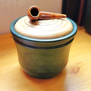 May include: A ceramic jar with a deep green base and a light tan lid. The lid features a circular, spiral-like texture and is topped with a small, brown, pipe-shaped handle. The jar has a smooth, rounded body and a wide, dark green rim.