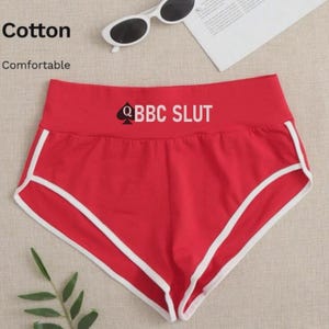 May include: Red shorts with white trim and the text "Q BBC SLUT". The shorts are made of cotton and are described as comfortable. Sunglasses and a magazine are in the background.