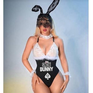 Könnte beinhalten: Ein schwarz-weißes Snow Bunny Kostüm-Set. Das Set beinhaltet einen Body mit dem Text "Snow Bunny" und einem Pik-Symbol, einen Spitzen-Hasenohren-Kopfschmuck, einen Choker und Armbänder.