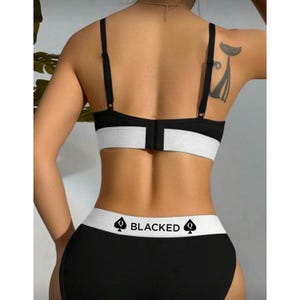 K&ouml;nnte beinhalten: Schwarzes und wei&szlig;es Dessous-Set. Der BH und die Unterw&auml;sche haben eine schwarze Basis mit einem wei&szlig;en Band. Die Unterw&auml;sche hat das Wort "BLACKED" auf dem Bund aufgedruckt, zusammen mit einem Pik-Symbol.