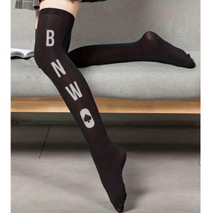 Puede incluir: Medias negras hasta el muslo con texto blanco "BNWO" y un símbolo de pica blanco. Las medias están hechas de un material transparente y se usan en las piernas.