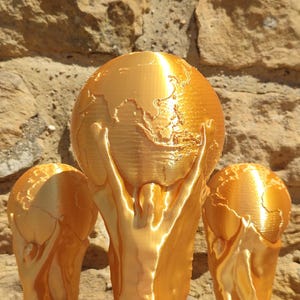 Puede incluir: Un trofeo dorado con un dise&ntilde;o de globo terr&aacute;queo sostenido por dos figuras. El trofeo est&aacute; sobre un muro de piedra texturizado. El trofeo es un s&iacute;mbolo de logro.