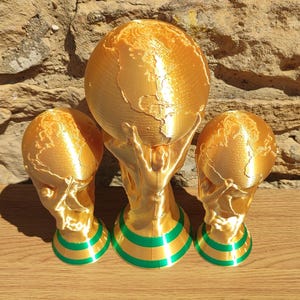 Puede incluir: Tres esculturas de trofeos dorados con un dise&ntilde;o de globo terr&aacute;queo, sostenidas por manos, y dos trofeos m&aacute;s peque&ntilde;os a juego. Las bases tienen rayas verdes y doradas. Los trofeos se exhiben sobre una superficie de madera contra una pared de piedra.