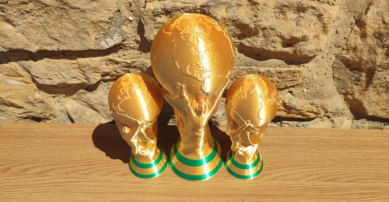 Puede incluir: Tres esculturas de trofeos dorados con un dise&ntilde;o de globo terr&aacute;queo, sostenidas por manos, y dos trofeos m&aacute;s peque&ntilde;os a juego. Las bases tienen rayas verdes y doradas. Los trofeos se exhiben sobre una superficie de madera contra una pared de piedra.