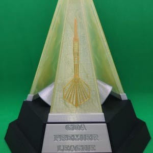 May include: A trophy with a pyramid-shaped top and a dart design inside. The base is black with the text "GDA PREMIER LEAGUE". The trophy is set against a green background.
