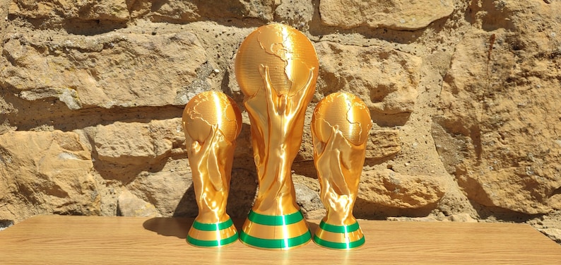 Puede incluir: Tres trofeos dorados con un dise&ntilde;o de globo terr&aacute;queo y figuras sosteniendo el mundo. Los trofeos tienen bases a rayas verdes y doradas. Los trofeos se exhiben sobre una superficie de madera contra una pared de piedra.