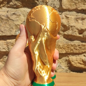 Puede incluir: Un trofeo dorado con una parte superior en forma de globo y una base verde y dorada. El trofeo se sostiene en una mano sobre un fondo de piedra texturizada. El trofeo es una representaci&oacute;n del mundo.