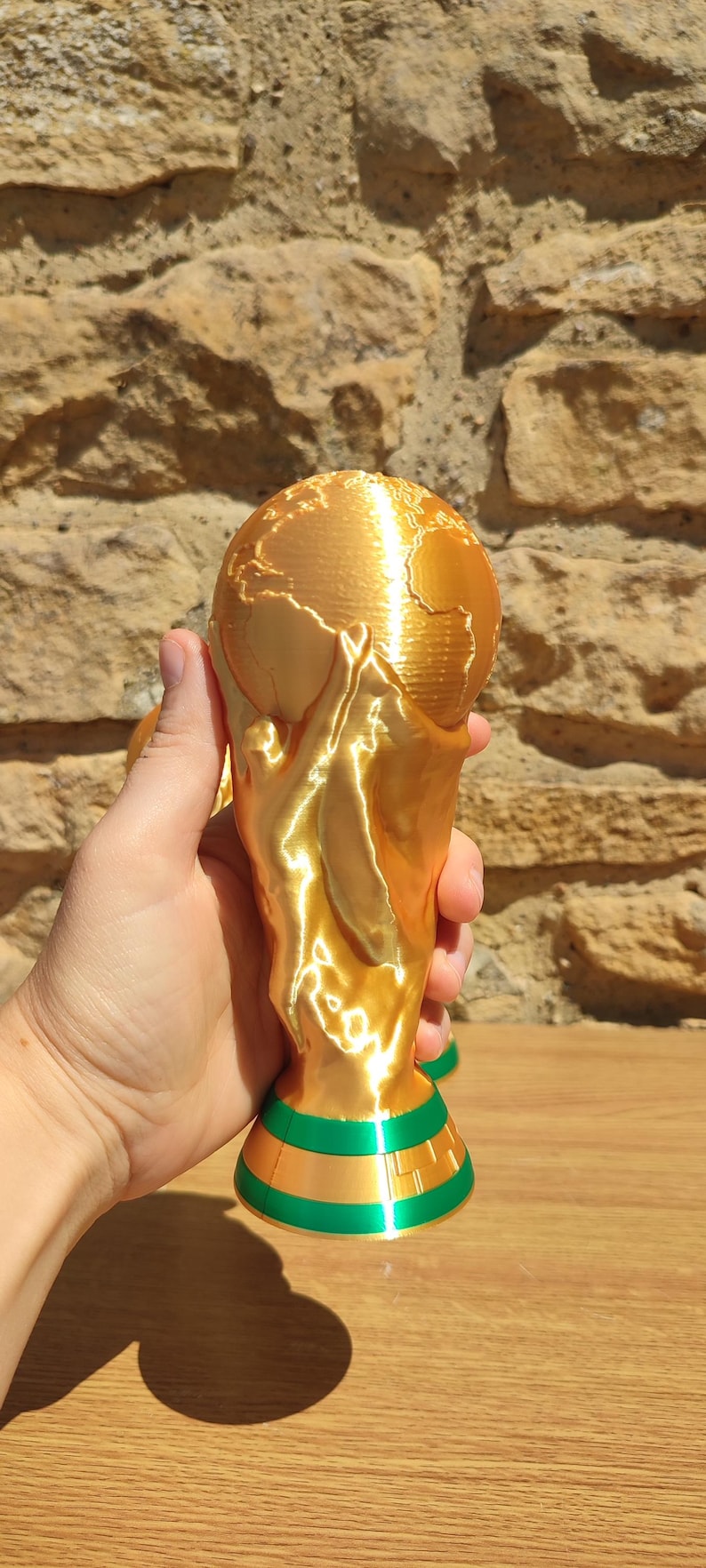Puede incluir: Un trofeo dorado con una parte superior en forma de globo y una base verde y dorada. El trofeo se sostiene en una mano sobre un fondo de piedra texturizada. El trofeo es una representaci&oacute;n del mundo.