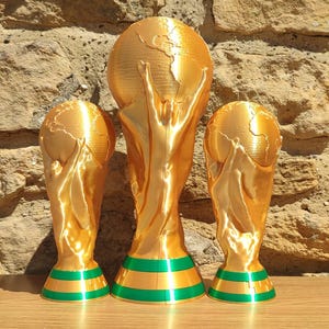 Puede incluir: Tres estatuas de trofeos dorados con bases a rayas verdes y doradas. Los trofeos tienen la forma de un globo terr&aacute;queo sostenido por dos figuras. Los trofeos se exhiben sobre una superficie de madera contra una pared de piedra.
