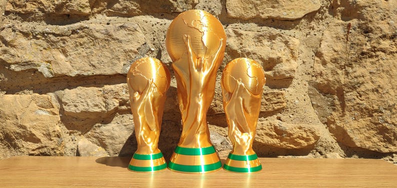 Puede incluir: Tres estatuas de trofeos dorados con bases a rayas verdes y doradas. Los trofeos tienen la forma de un globo terr&aacute;queo sostenido por dos figuras. Los trofeos se exhiben sobre una superficie de madera contra una pared de piedra.