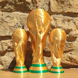 Puede incluir: Tres r&eacute;plicas de trofeos dorados con bases verdes y doradas. Los trofeos son de diferentes tama&ntilde;os, el m&aacute;s grande en el centro. Los trofeos tienen un globo terr&aacute;queo en la parte superior y figuras que lo sostienen.