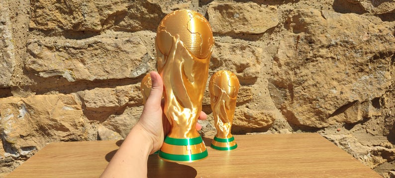 Puede incluir: Dos r&eacute;plicas de trofeos dorados con bases verdes y doradas. El trofeo m&aacute;s grande est&aacute; sostenido por una mano. Los trofeos est&aacute;n sobre una superficie de madera frente a una pared de piedra. Los trofeos est&aacute;n detallados con dise&ntilde;os de globo y figuras.