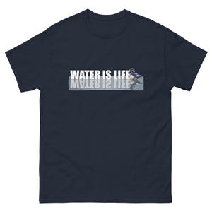 Peut inclure: Un t-shirt bleu marine à manches courtes avec le texte blanc WATER IS LIFE imprimé sur la poitrine. À droite du texte se trouve un petit graphique représentant un canard nageant, avec un reflet inversé du texte et du canard en dessous.