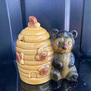 May include: A ceramic honey pot and bear figurine. The honey pot is shaped like a beehive with a lid and is painted yellow with brown accents. A brown bear figurine sits next to the honey pot, with a pink nose and blue eyes.