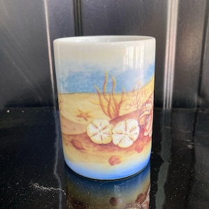 May include: A cylindrical ceramic tumbler with a coastal design. The artwork depicts a sandy beach with seashells, starfish, and aquatic plants under a blue sky. The base of the tumbler is a gradient of blue.