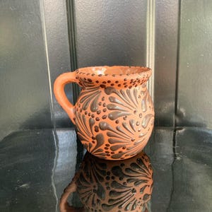 May include: A small, terracotta-colored mug with a handle. The mug is decorated with dark brown, stylized floral patterns and small dots. The rim of the mug is also dotted. The mug is sitting on a reflective surface.