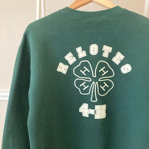 May include: A dark green sweatshirt with the word "HELOTES" arched above a white clover design with four "H" symbols. Below the clover is the text "4-H" in white. The sweatshirt is hanging on a wooden hanger.