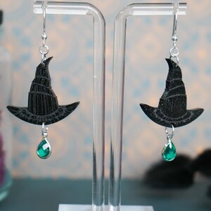 Puede incluir: Un par de pendientes de sombrero de bruja negros con una gema verde en forma de lágrima. Los pendientes se exhiben en un soporte de acrílico transparente. Los sombreros de bruja tienen un diseño texturizado y están suspendidos de ganchos plateados.