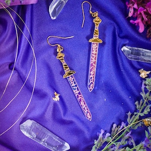 Puede incluir: Un par de pendientes dorados con forma de espada, con detalles en morado y azul, sobre un fondo de satén morado. Los pendientes presentan diseños intrincados y están acompañados de cristales de cuarzo y ramitas de lavanda.