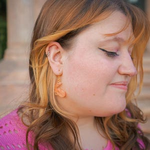 May include: Close-up of a person wearing gold hoop earrings with peach-colored, abstract-shaped dangles. The person is wearing a pink sweater. The background is blurred, with a building visible.