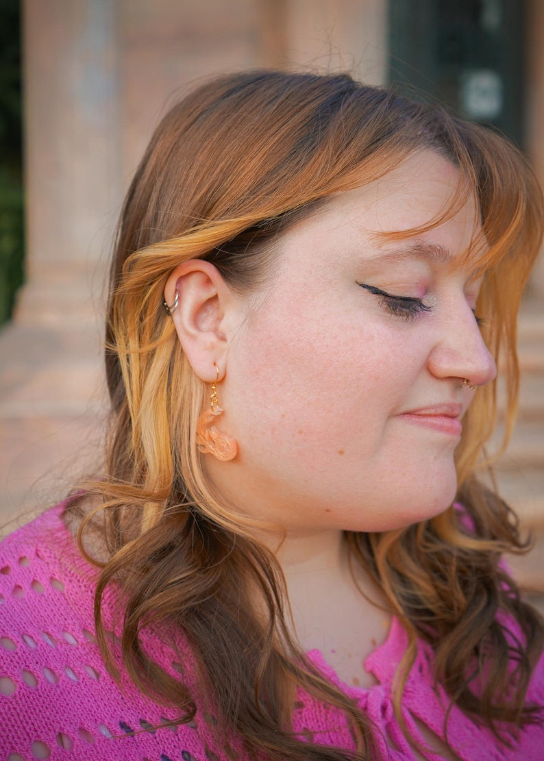 May include: Close-up of a person wearing gold hoop earrings with peach-colored, abstract-shaped dangles. The person is wearing a pink sweater. The background is blurred, with a building visible.