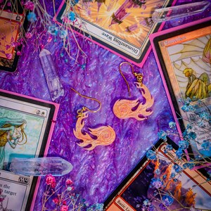 May include: A pair of flame-shaped earrings with gold-colored hooks, set against a purple and pink background with floral accents and collectible cards. The earrings are a light orange color.