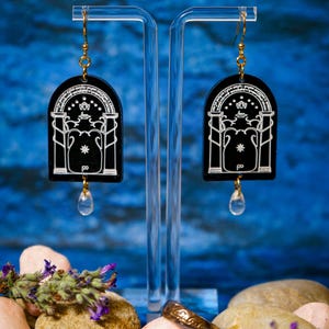 May include: Black acrylic earrings with a white design featuring an arched shape with a star and floral details. The earrings hang from gold-colored hooks and have a clear teardrop-shaped charm. A gold ring and stones are in the foreground.
