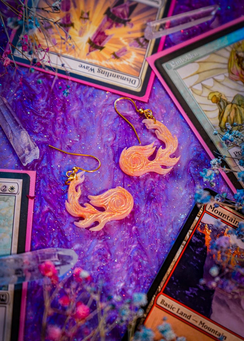 May include: A pair of flame-shaped earrings with gold-colored hooks. The earrings are a light orange color with a swirling design. The background is a purple, glittery surface with collectible cards and crystals.