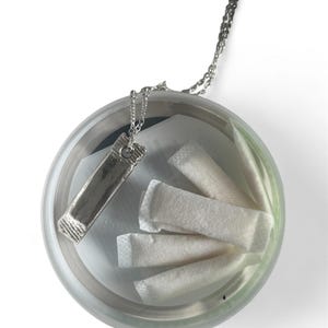 Puede incluir: Un collar plateado con un colgante rectangular descansa dentro de un recipiente redondo y transparente. El recipiente contiene varios paquetes blancos y rectangulares. La cadena del collar es visible, saliendo del recipiente.