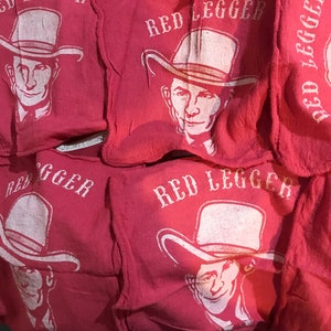 Puede incluir: Una pila de cuadrados de tela roja con una impresión blanca de un hombre con un sombrero y el texto "RED LEGGER".