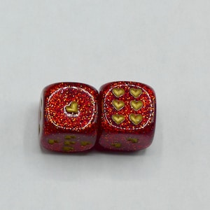 May include: Two red glitter dice with gold heart pips. One die shows a single heart, the other shows six hearts. The dice are set against a white background.