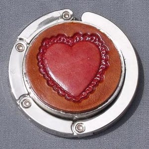 May include: A circular silver-toned metal bag hanger featuring a central inlay of brown leather. The leather insert is decorated with a raised, glossy red heart shape surrounded by a scalloped, embossed border detail within the circular frame.