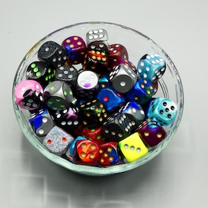 May include: A clear glass bowl filled with a variety of colorful dice. The dice are in various colors, including black, blue, pink, and yellow, with white or colored pips. The dice are of standard size, suitable for tabletop games.