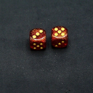 May include: Two red dice with gold dots. The dice are on a black surface.